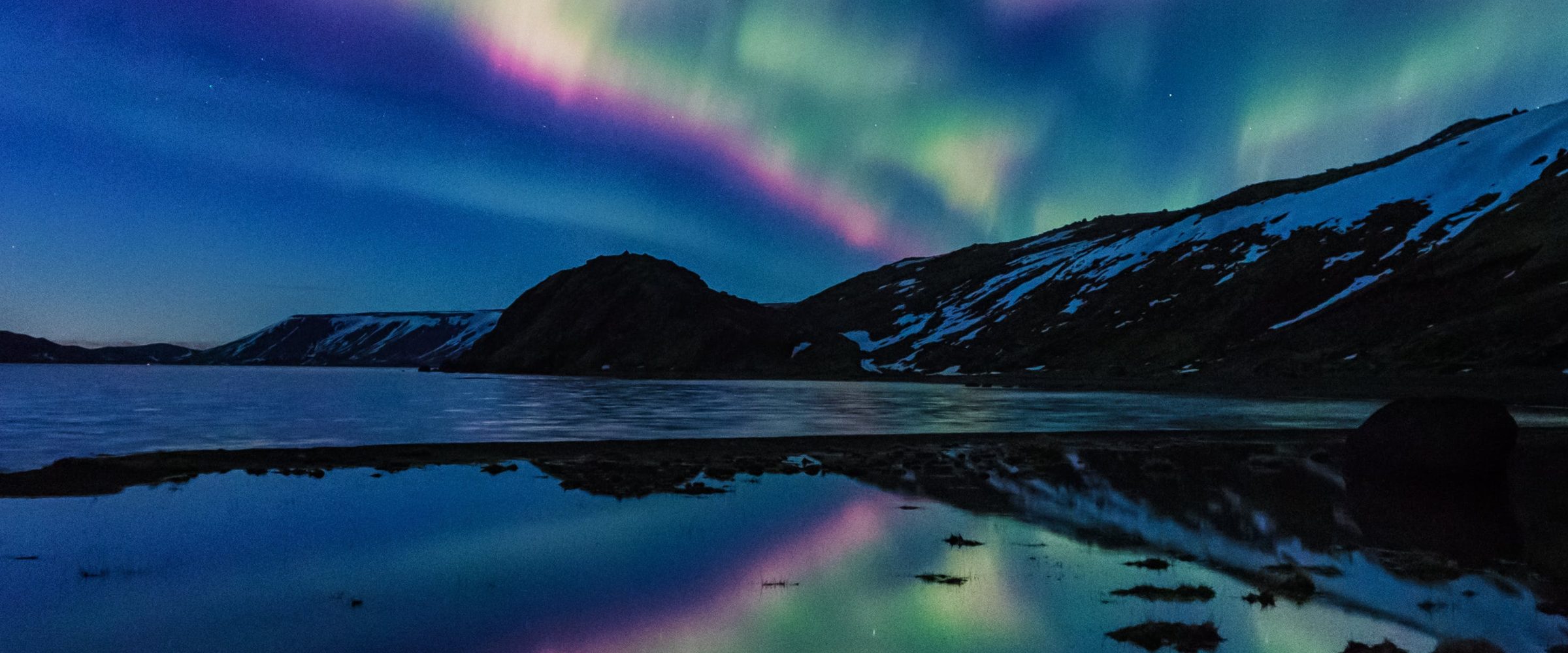 The Northern Lights over a still lake and snow-patched fells in Finnish Lapland